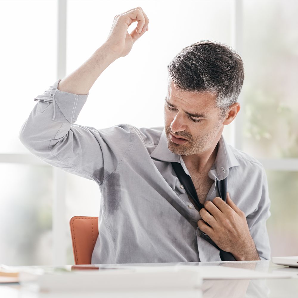 Mann im Hemd, Arm unter Achsel. Schweißflecken sichtbar. Er zieht Krawatte runter.