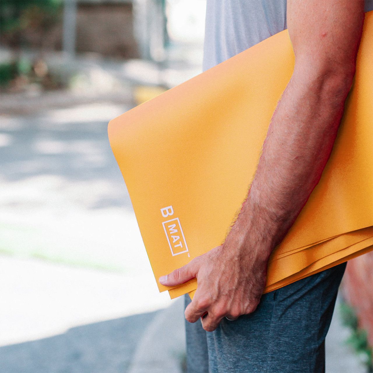 Person hält eine gefaltete, gelb-orange Yogamatte. Logo B MAT sichtbar.