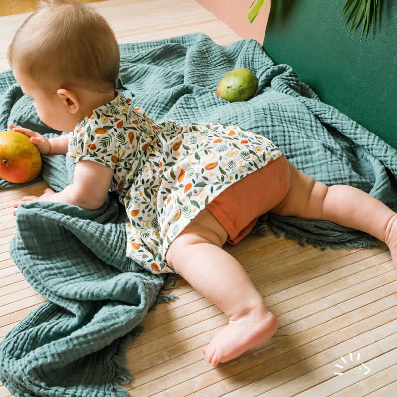 Baby trägt Bio-BW Mango Kleid. Liegt auf Decke, hält Mango. Kleid mit Fruchtmuster, orangefarbener Unterteil. Kurze Ärmel.