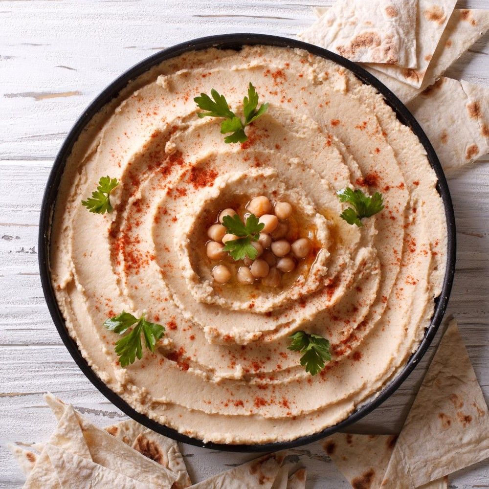 Hummus in schwarzer Schüssel, spiralförmig angerichtet. Mit Kichererbsen, Öl und Paprika bestreut. Petersilie als Garnierung. Fladenbrotstücke.