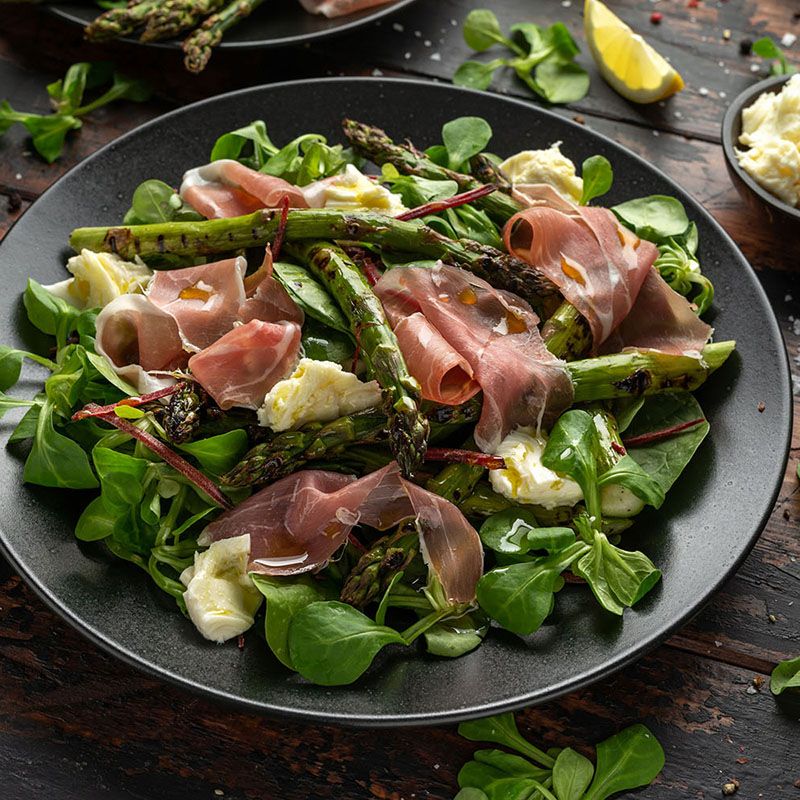 Schwarzer Teller mit Salat, Spargel, Schinken und Käse. Grüner Salat, Spargelstangen, Schinkenscheiben und Käsewürfel.