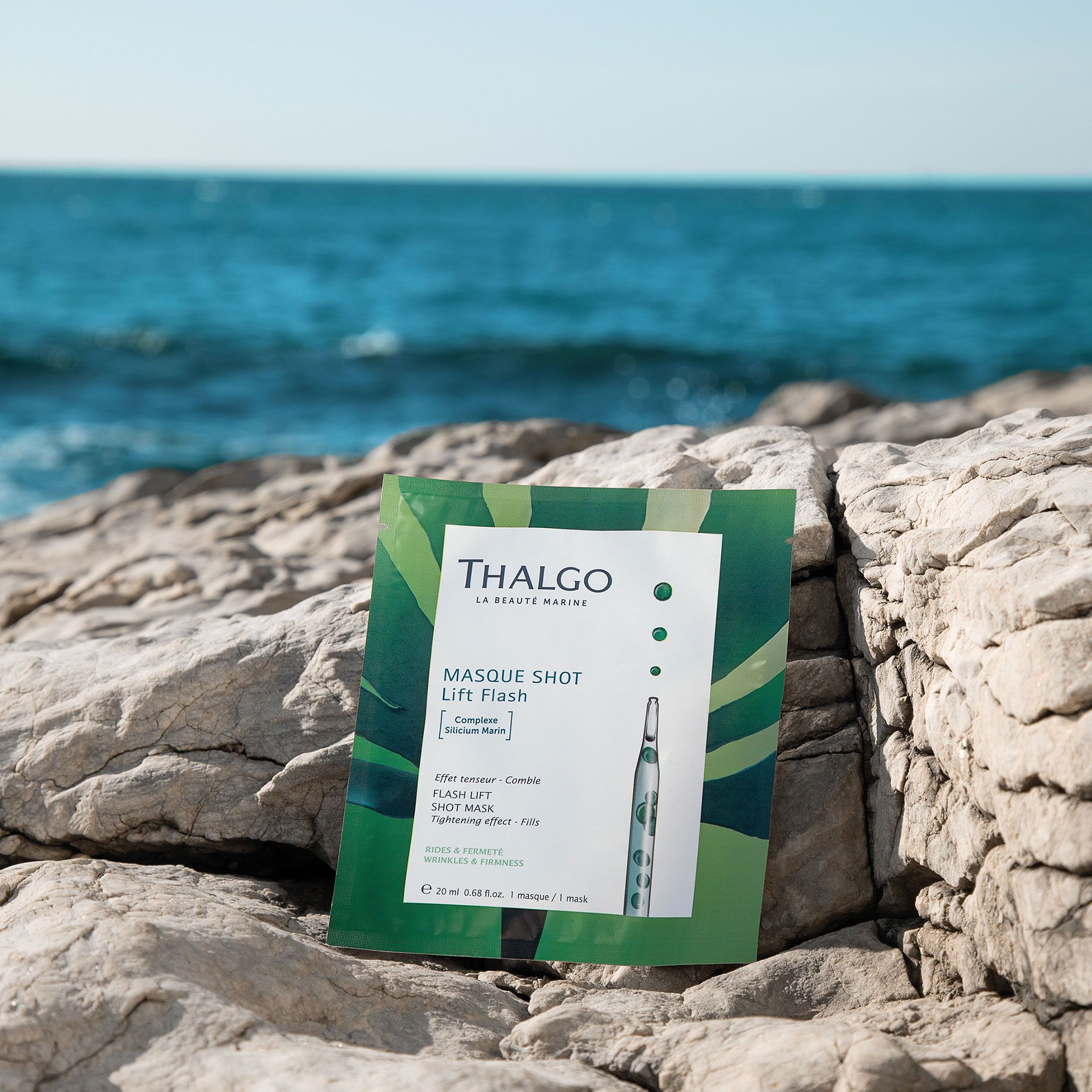 Grüne Einzelverpackung auf Felsen am Meer. Produktname und Logo sichtbar. Enthält eine Ampulle. Text: Masque Shot Lift Flash.