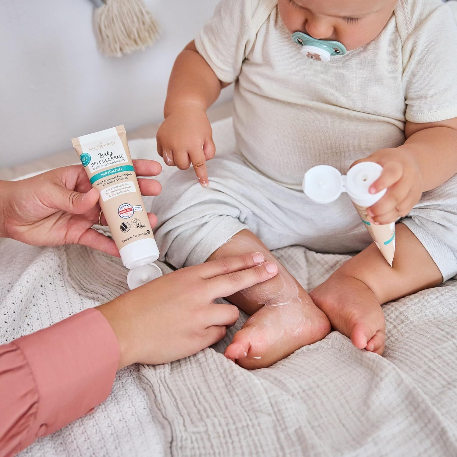 Ein Baby, dem Creme auf das Bein aufgetragen wird. Eine Person hält eine Tube MABYEN Baby Pflegecreme. Creme wird auf das Bein des Babys aufgetragen.