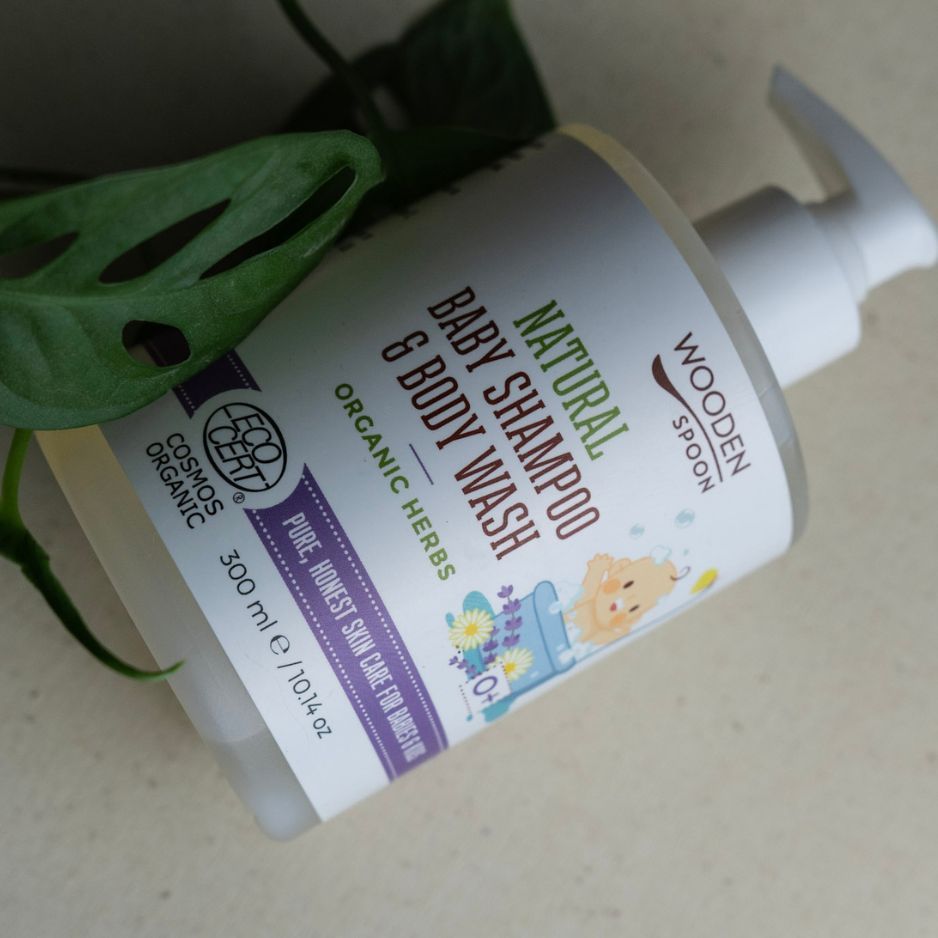 Baby-Shampoo- und Body Wash-Flasche. Weißer Spender, Etikett mit Text und Illustrationen. Cosmos Organic-Zertifizierung. Teilweise verdeckt von Blatt.