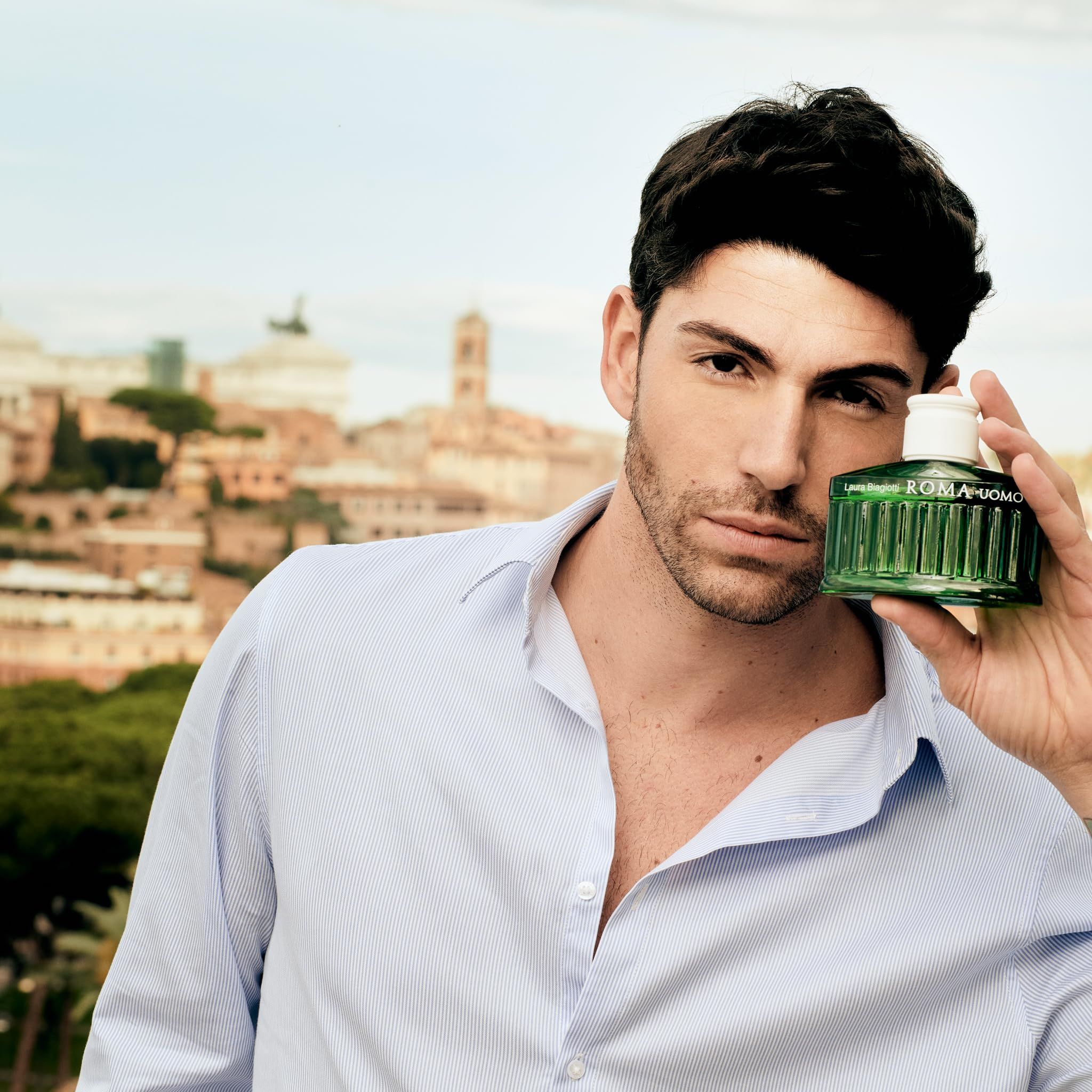 Mann hält grüne Flasche. Im Hintergrund Stadtansicht. Flasche mit Aufschrift: ROMA UOMO.