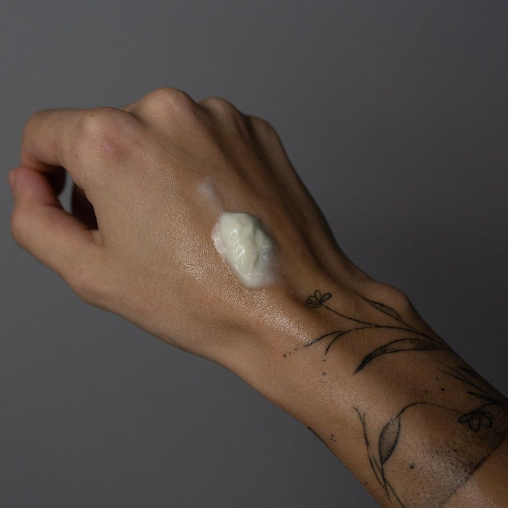 Hand mit Tattoo, auf der eine kleine Menge Creme liegt. Grauer Hintergrund.