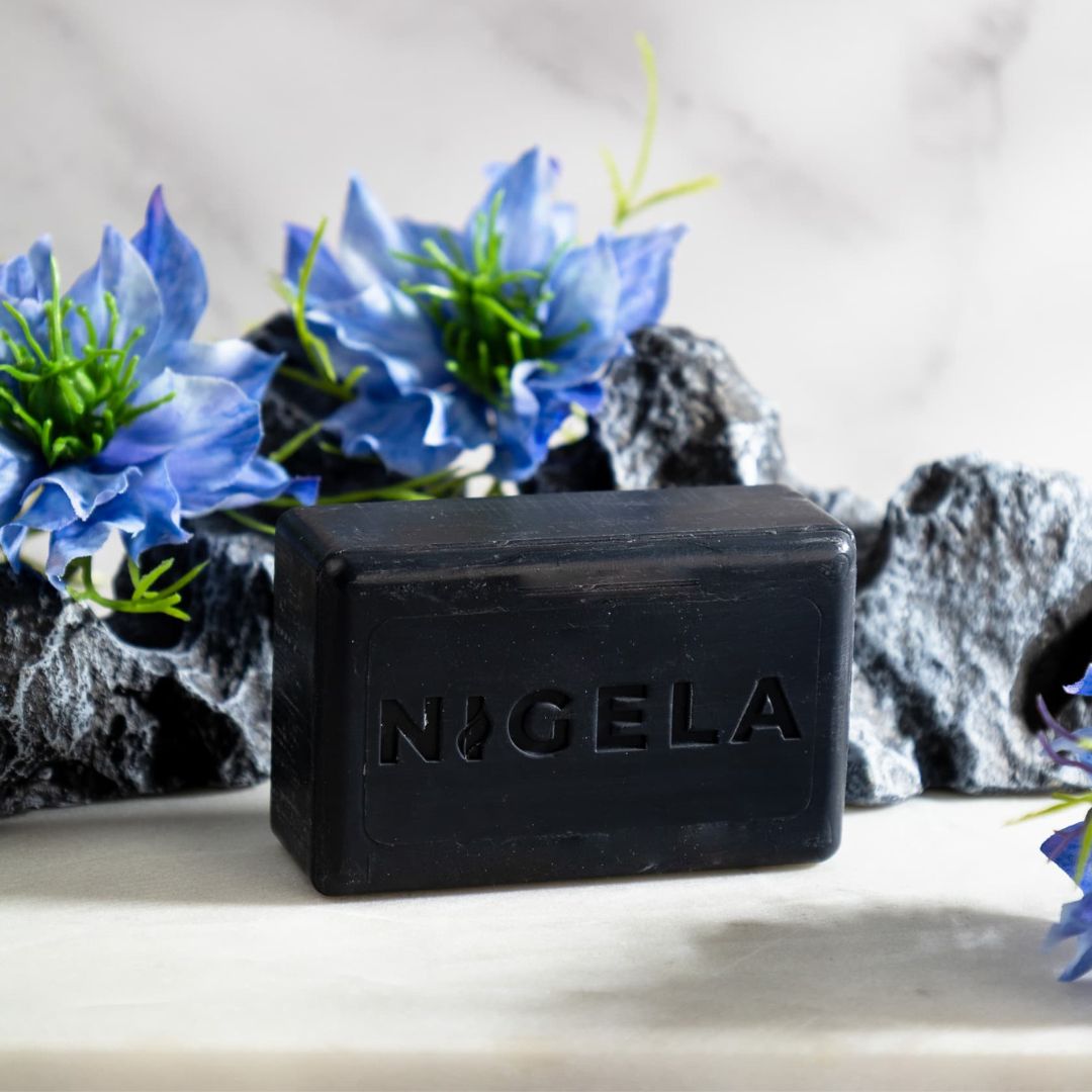 Schwarze Seife mit geprägtem Nigela-Logo. Rechteckige Form. Dekoriert mit blauen Blüten und Steinen.
