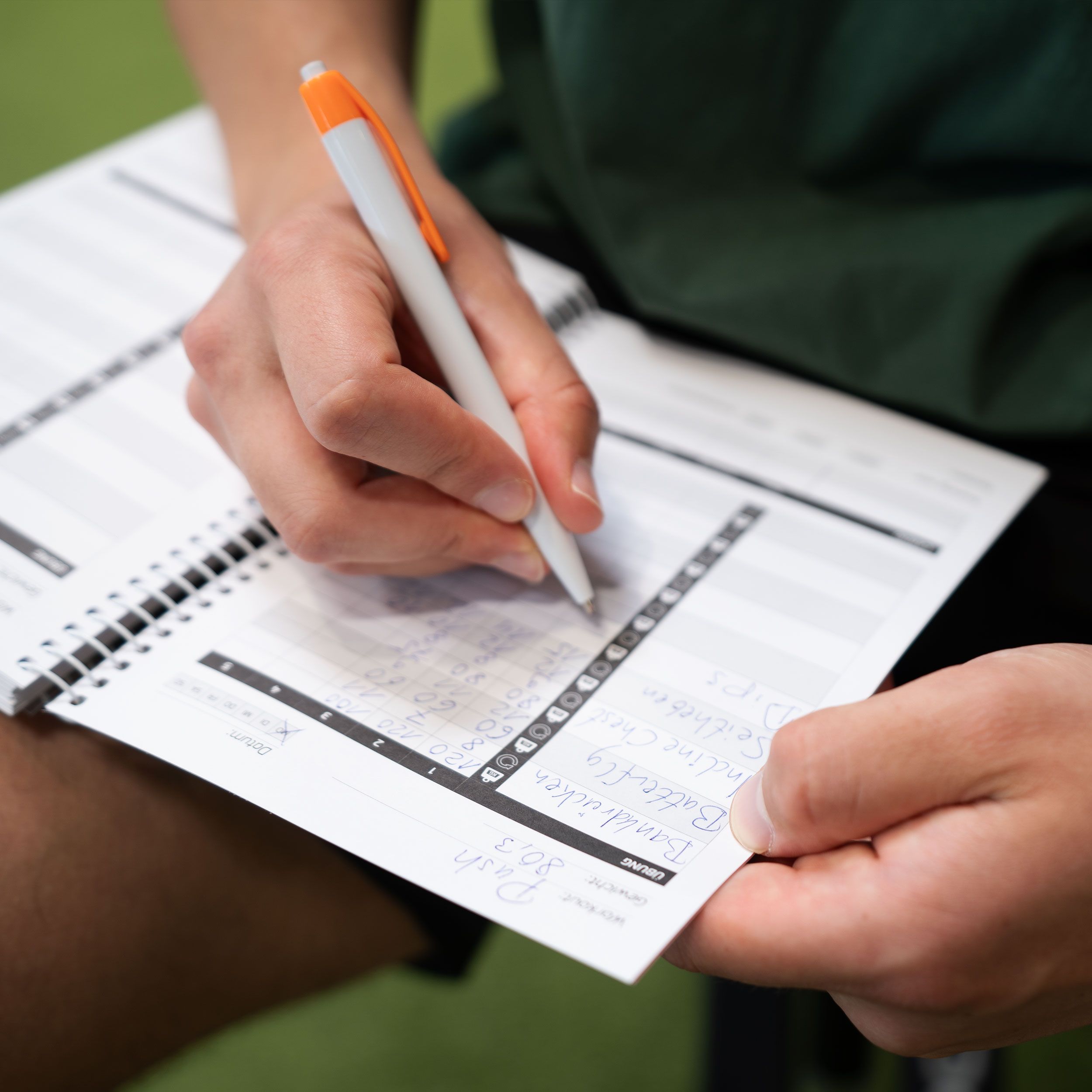 Person schreibt in ein offenes Trainingsbuch. Buch mit Spiralbindung. Tabellen und Diagramme auf den Seiten. Stift in der Hand.