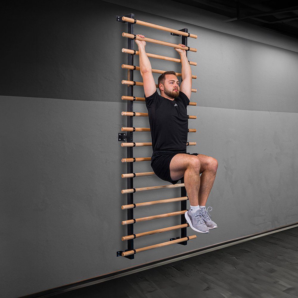 Mann an Sprossenwand. Er sitzt und hält sich an den Sprossen fest. Schwarzer Rahmen, Holzsprossen.