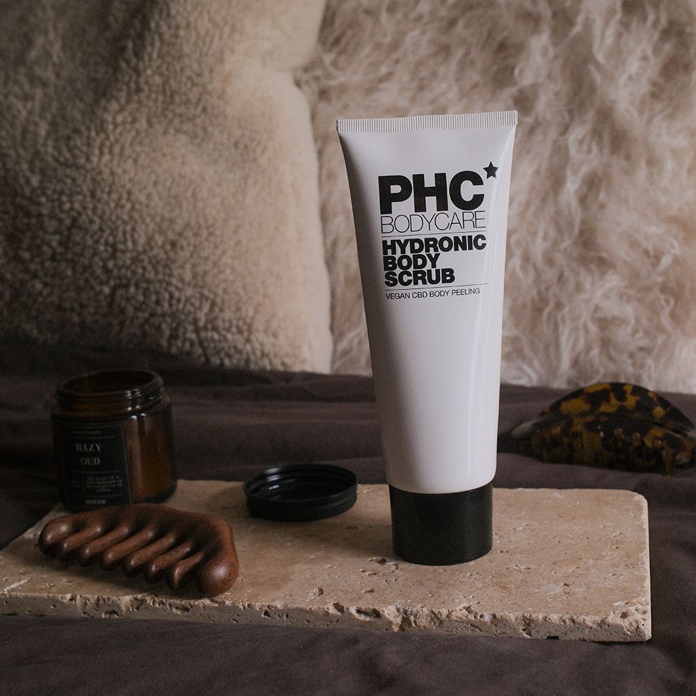 Weiße Tube PHC Bodycare Hydronic Body Scrub auf Steinplatte. Schwarzer Deckel, Holzmassagegerät und Glasbehälter daneben. Vegan CBD Body Peeling.
