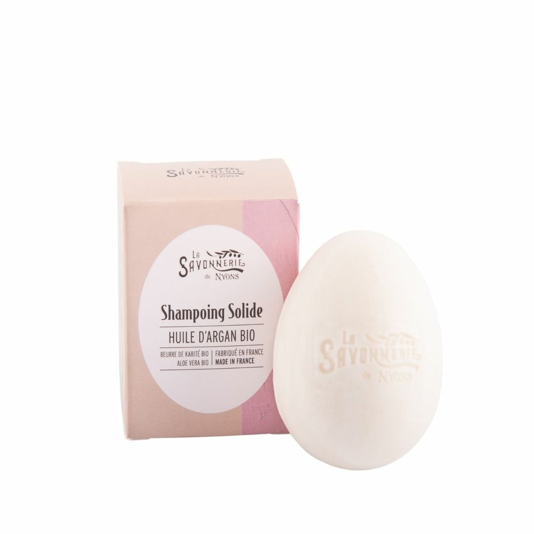Eiförmiges, festes Shampoo neben Kartonverpackung. Rosa-beige Farbverlauf. Weißes Etikett mit Text: Huile d'Argan Bio, Made in France.