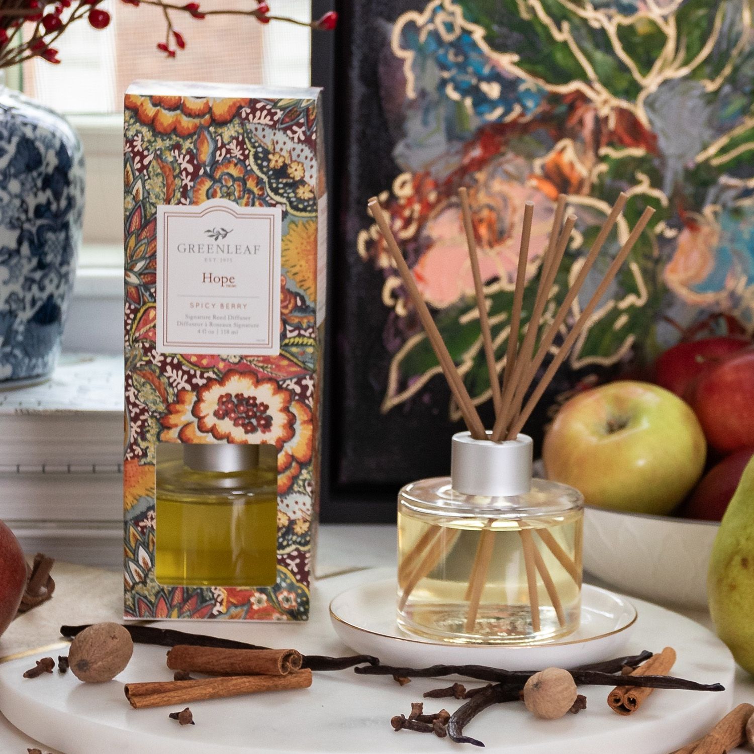 Greenleaf Hope Reed Diffuser mit 8 Stäbchen auf einem Teller, neben einer bunten Verpackung. Gewürze und Äpfel im Hintergrund.