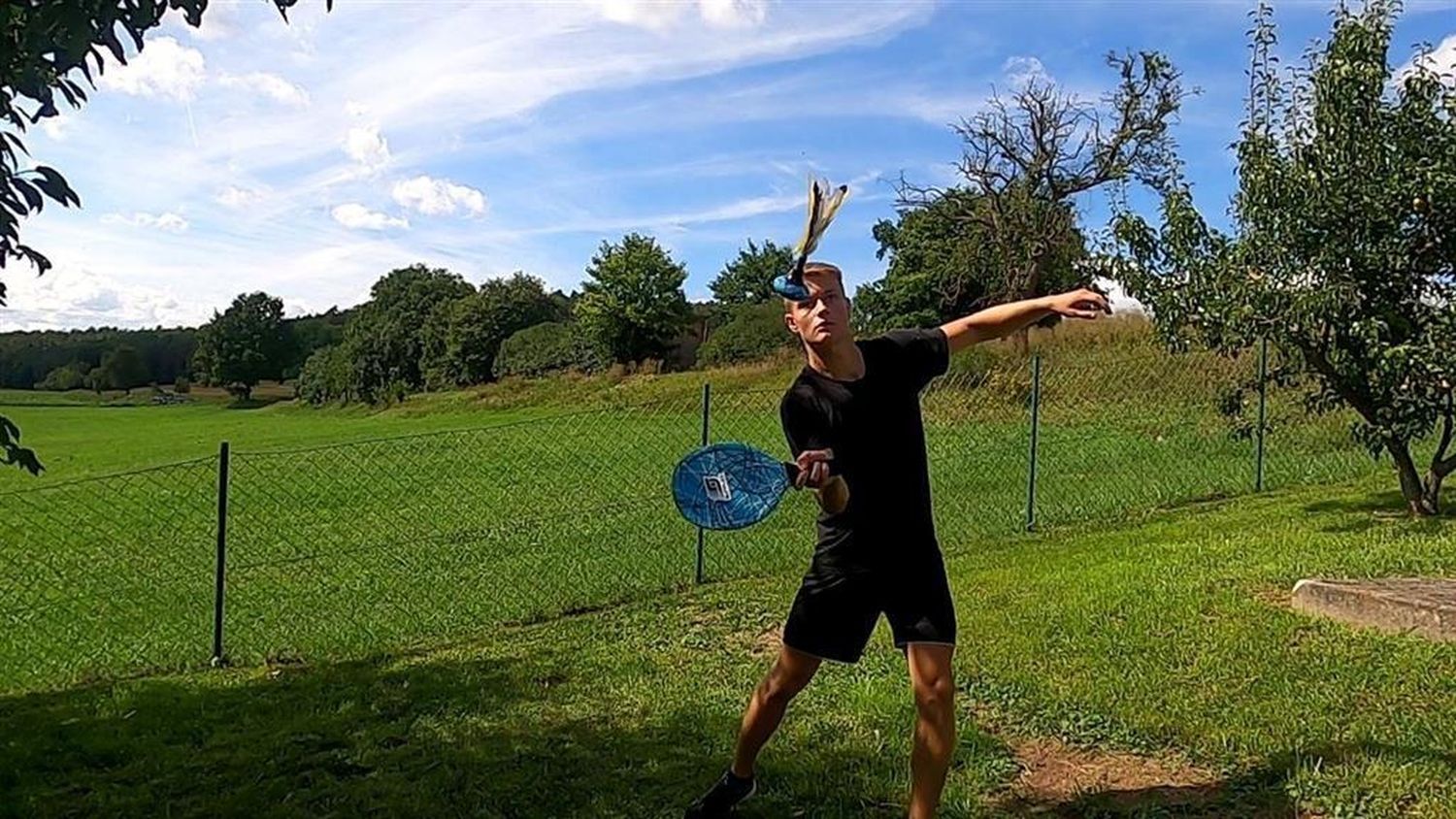 Mann spielt Peteca Neoremi Circle im Freien. Er trägt schwarze Kleidung und eine Sonnenbrille. Grüne Wiese im Hintergrund.