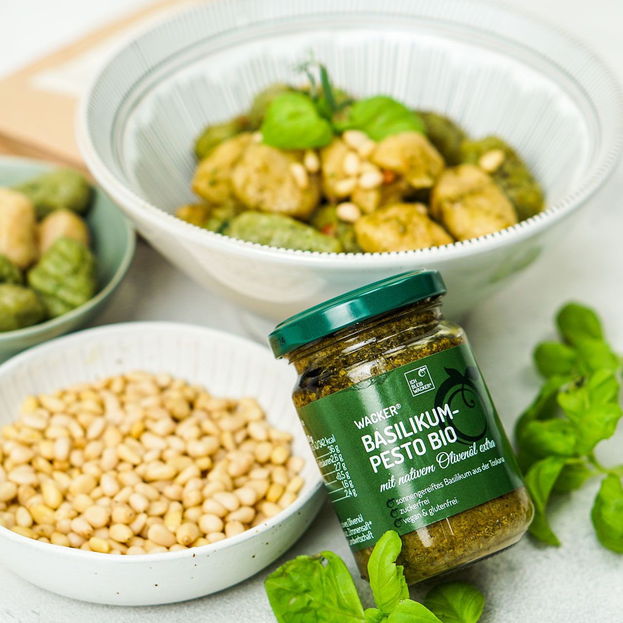 Glas Wacker Basilikumpesto Bio neben Schalen mit Gnocchi, Pinienkernen und Basilikumblättern. Grüner Deckel, grüne Beschriftung.