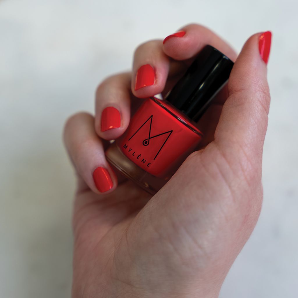Hand hält eine rote Nagellackflasche. Nägel sind rot lackiert. Auf der Flasche steht Mylène.