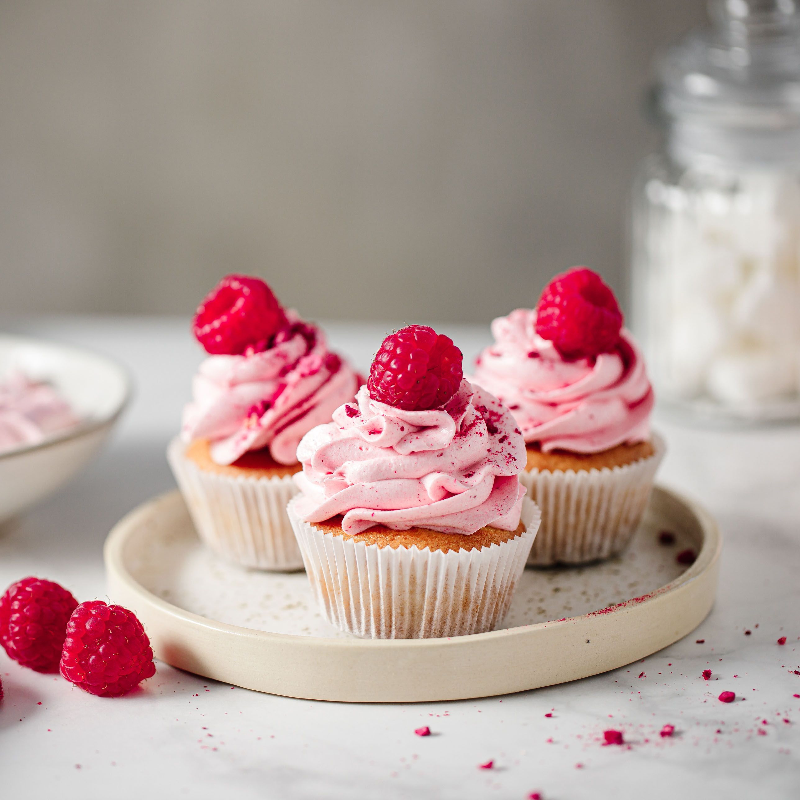 Drei Cupcakes mit rosa Creme und Himbeere. Auf einem Teller. Weitere Himbeeren daneben.