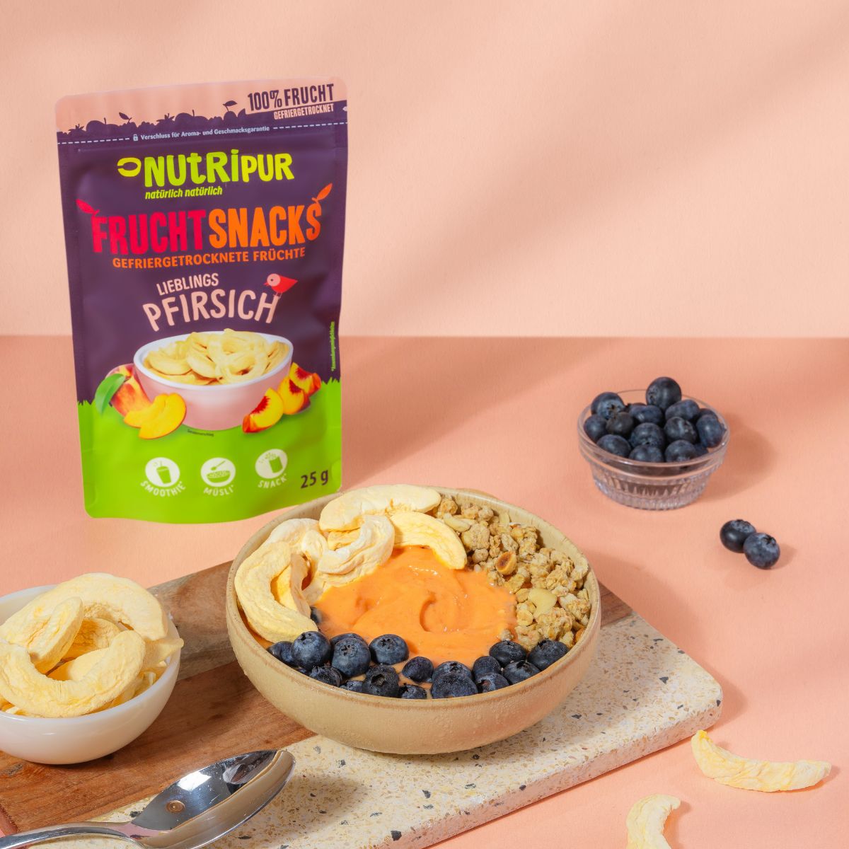 Schale mit Smoothie, Blaubeeren und gefriergetrockneten Pfirsich-Fruchtsnacks. Verpackung NutriPur.