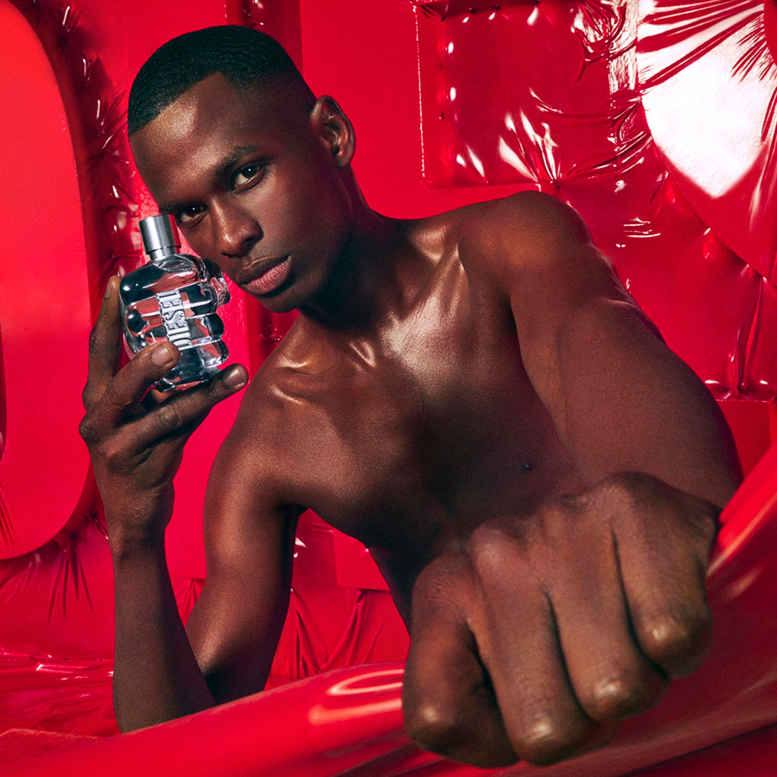 Mann hält Diesel Only The Brave Flakon. Flakon in Faustform, blaues Glas. Roter Hintergrund.