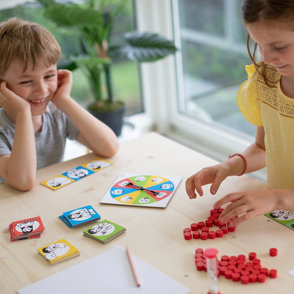 Zwei Kinder spielen am Tisch. Karten, Spielscheibe, Holzplättchen, Stift und Sanduhr sind sichtbar.