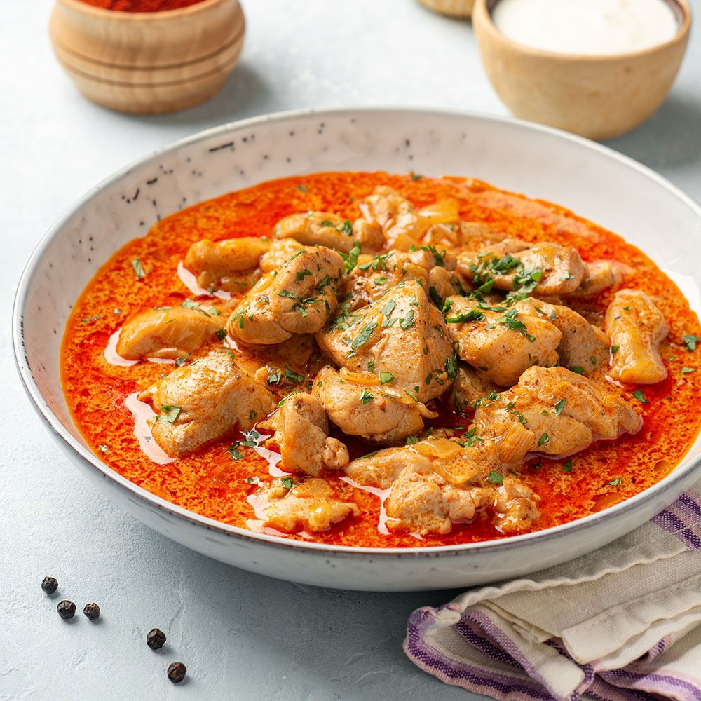 Hühnergericht in einer Schüssel. Hühnerstücke in roter Sauce. Garniert mit Kräutern. Schwarze Pfefferkörner.