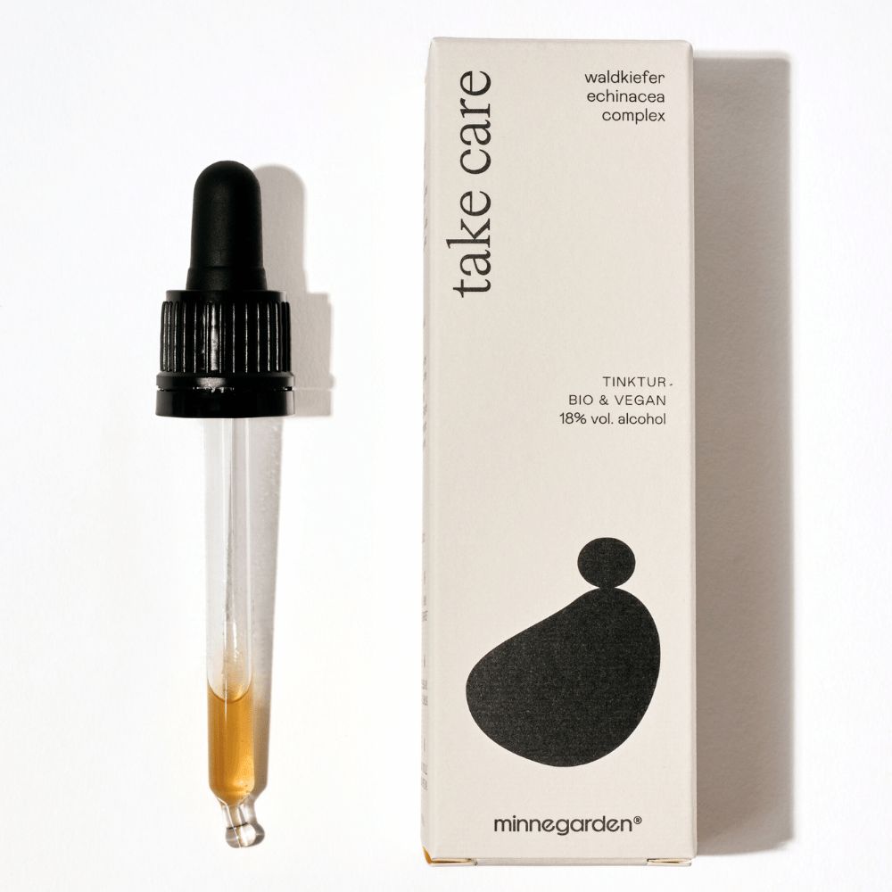 Braune Flüssigkeit in einer Pipette und eine weiße Schachtel mit schwarzer Schrift. Auf der Schachtel steht "take care" und "minnegarden".