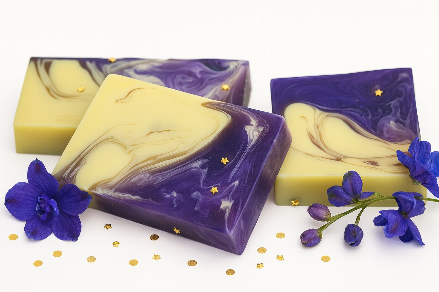 Drei Seifenstücke mit gelb-lila Marmorierung und goldenen Sternen. Blaue Blüten und goldene Konfetti liegen daneben.