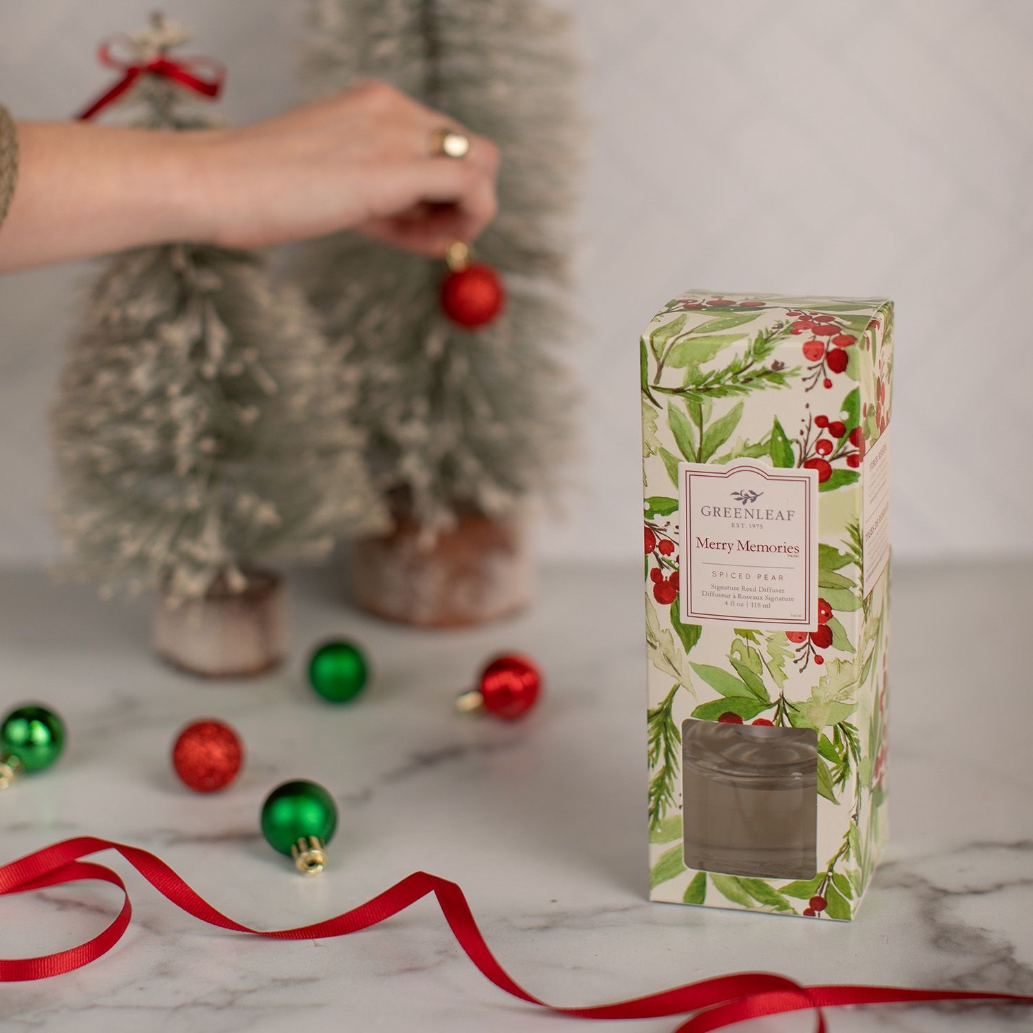 Duftspender mit Holzstäbchen und Verpackung neben kleinen Weihnachtsbäumen und Christbaumkugeln.