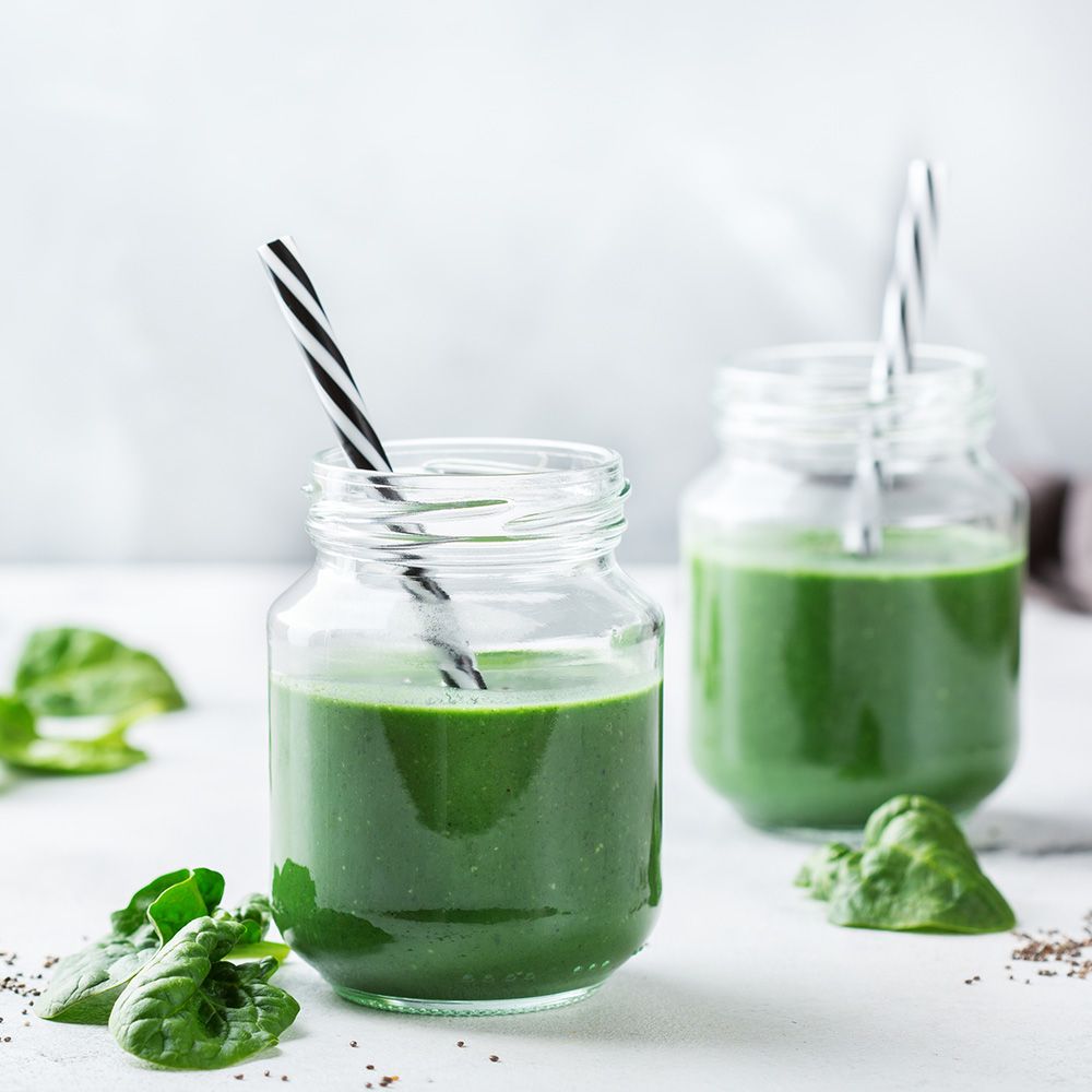 Zwei grüne Smoothies in Gläsern mit Strohhalmen. Blätter und Samen liegen daneben.