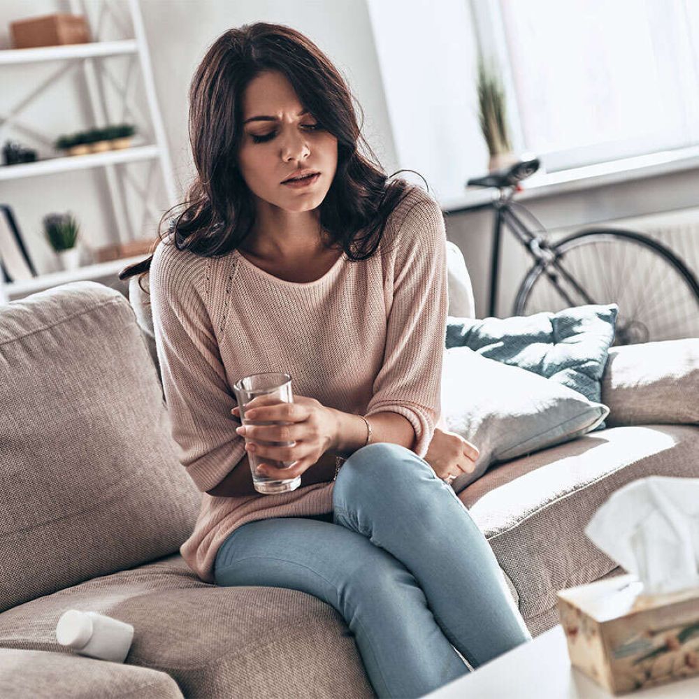 Frau sitzt auf Sofa, hält Glas Wasser, wirkt unwohl. Neben ihr Taschentücher und Medikamente.