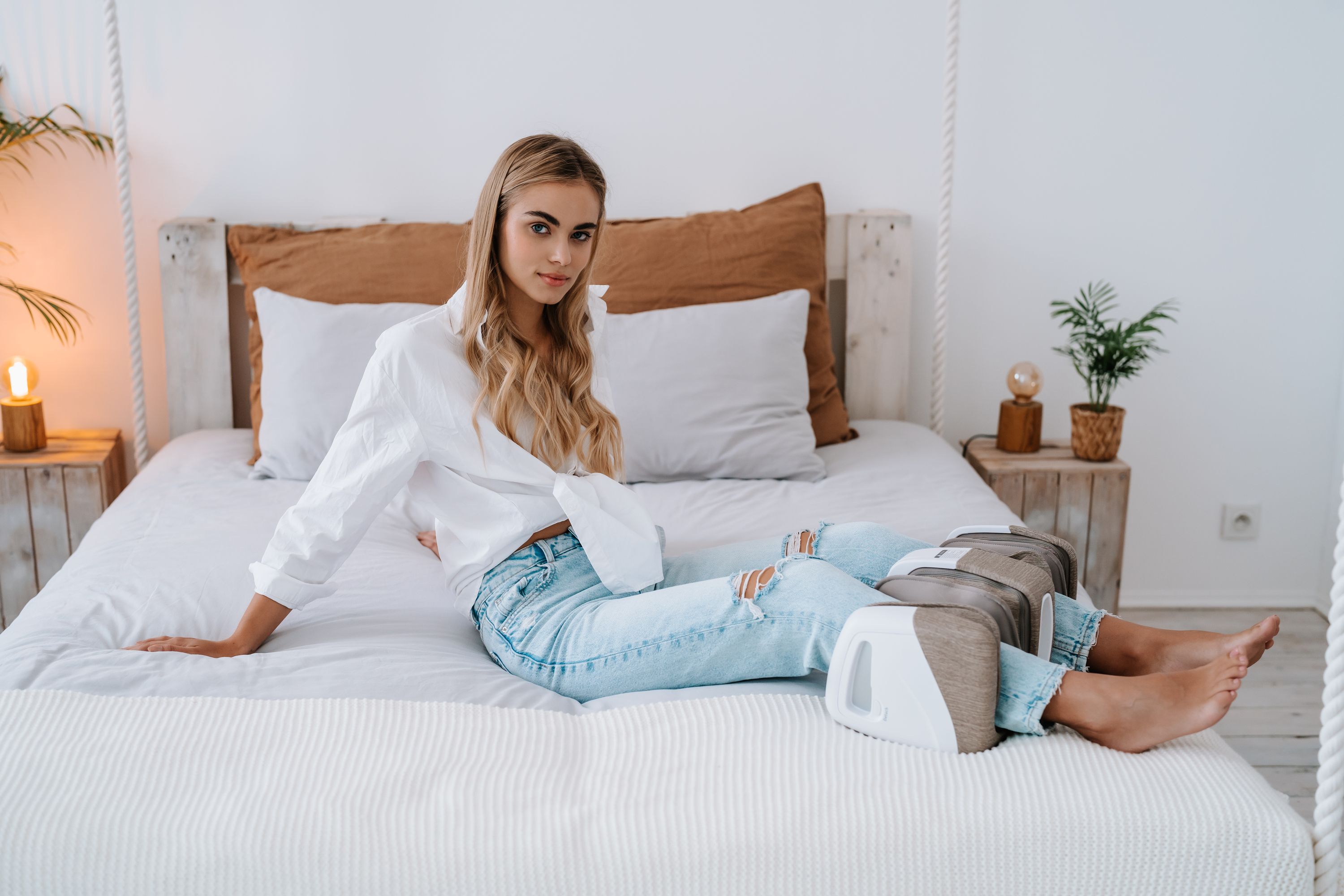Junge Frau sitzt auf einem Bett. Ein weißes Massagegerät ist neben ihr platziert. Sie trägt Jeans und ein weißes Hemd.