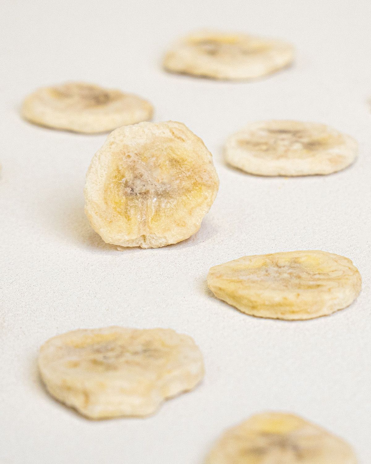 Nahaufnahme von gefriergetrockneten Bananenscheiben auf weißem Untergrund. Einzelne Scheiben verstreut.