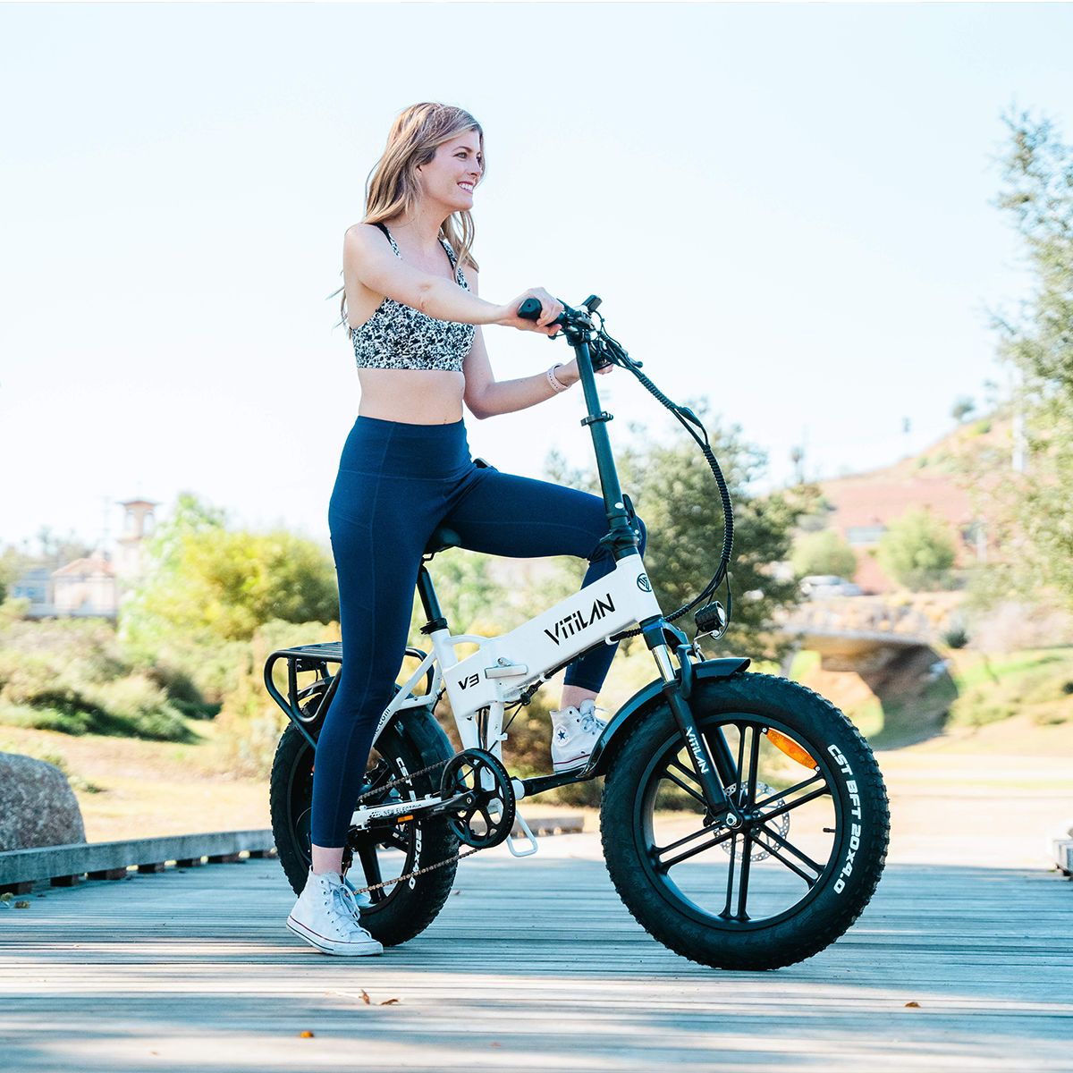 Weißes, zusammenklappbares E-Bike mit schwarzen Reifen. Frau fährt auf einer Brücke. Auf dem Rahmen steht Vitilan V3.