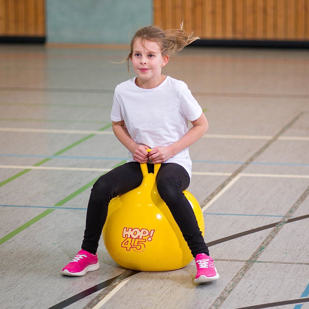Gelber Gymnic Hüpfball HOP! 45, Kind sitzt darauf. Ball mit Griff, Kind trägt Sportkleidung und Schuhe. In Sporthalle.