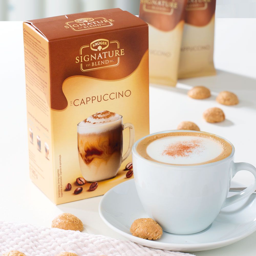 Karton mit Krüger Signature Blend Cappuccino. Cappuccino in Tasse auf Untertasse. Kekse daneben. Zwei Beutel im Hintergrund.