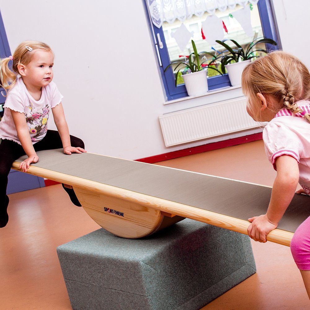 Balancebrett mit Klötzen und Riffelgummi. Zwei Kinder spielen damit. Sport-Thieme Logo sichtbar.