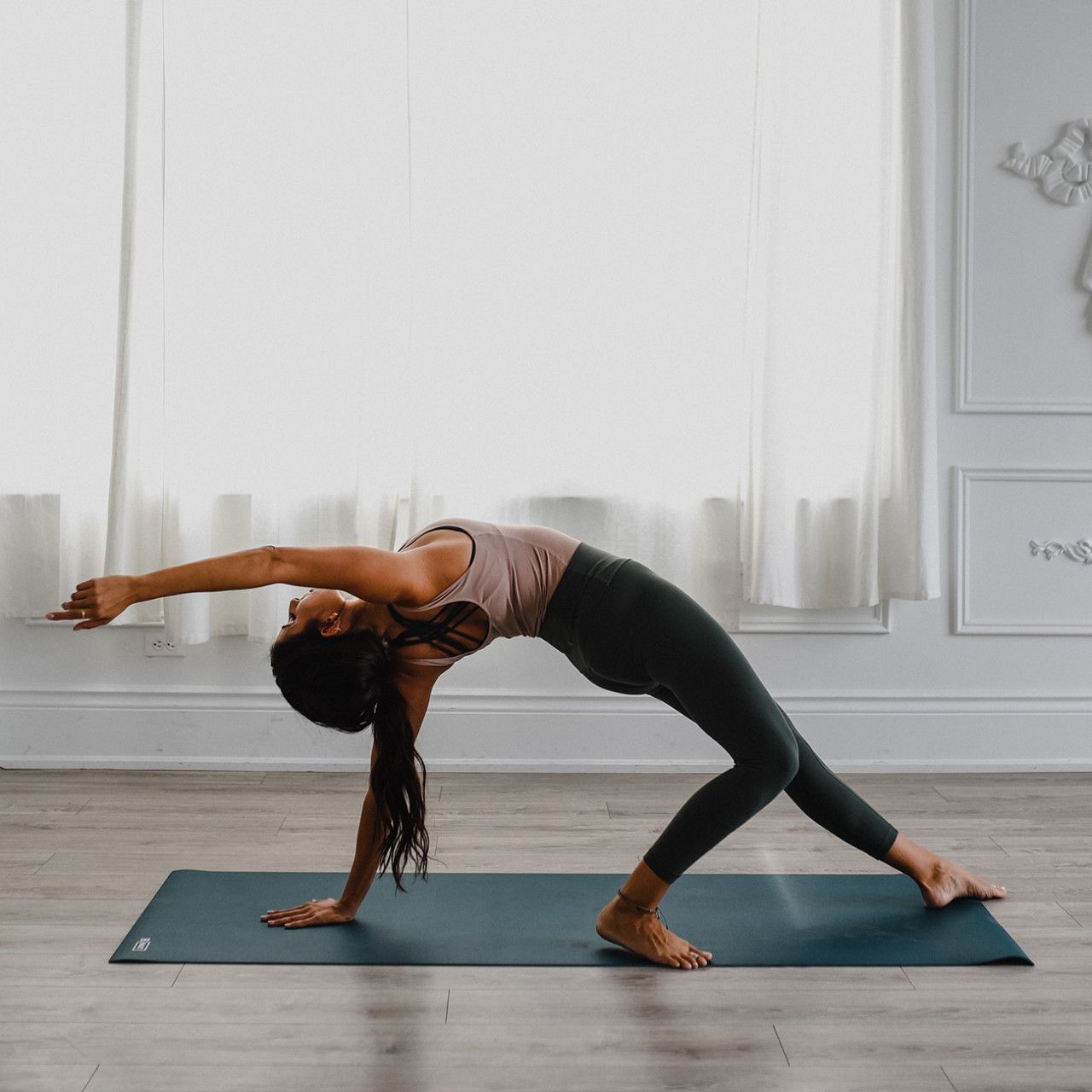 Person in Yoga-Pose auf dunkelgrüner Yogamatte. Hintergrund: Fenster, Vorhänge.