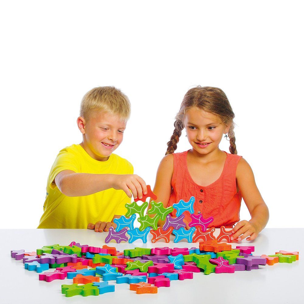 Zwei Kinder spielen mit bunten Holzfiguren. Ein Junge stapelt Figuren, ein Mädchen schaut zu. Figuren auf Tisch.