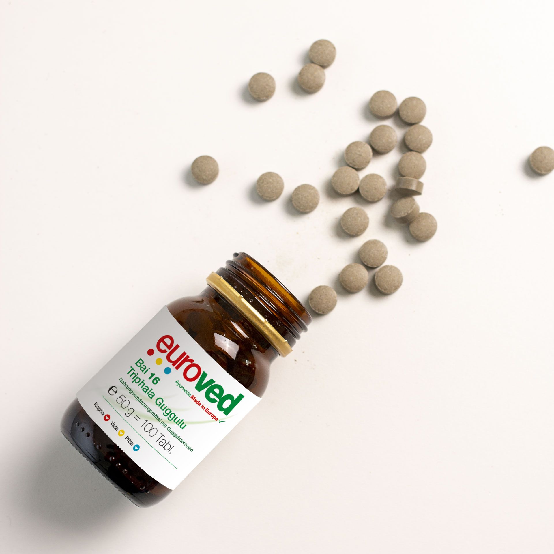 Braune Glasflasche mit Tabletten. Aufschrift: euroved, Bai 16 Triphala Guggulu. Tabletten liegen verstreut.