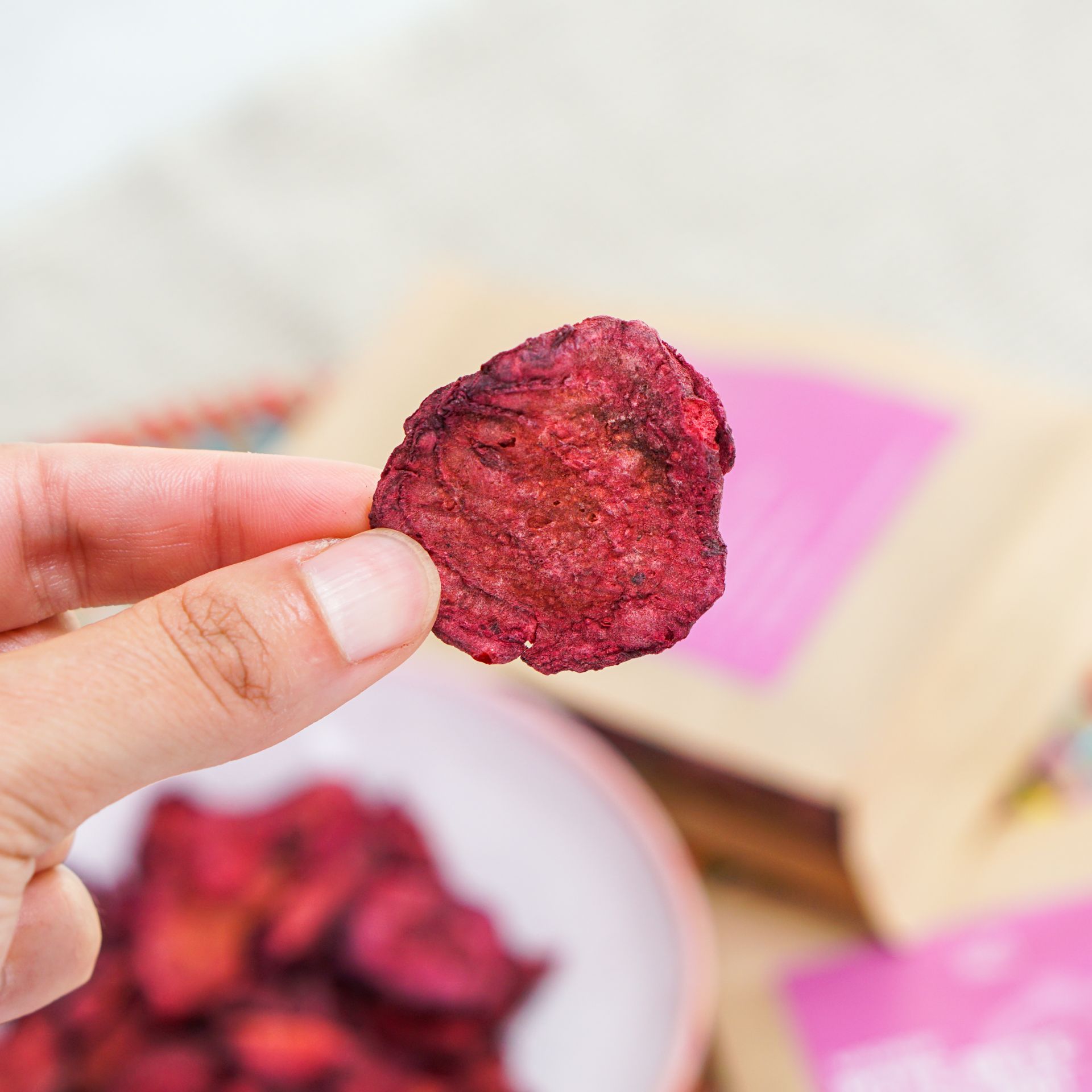 Hand hält einen Rote Bete Chip. Der Chip ist rot und unregelmäßig geformt. Im Hintergrund eine Schale und ein Beutel.