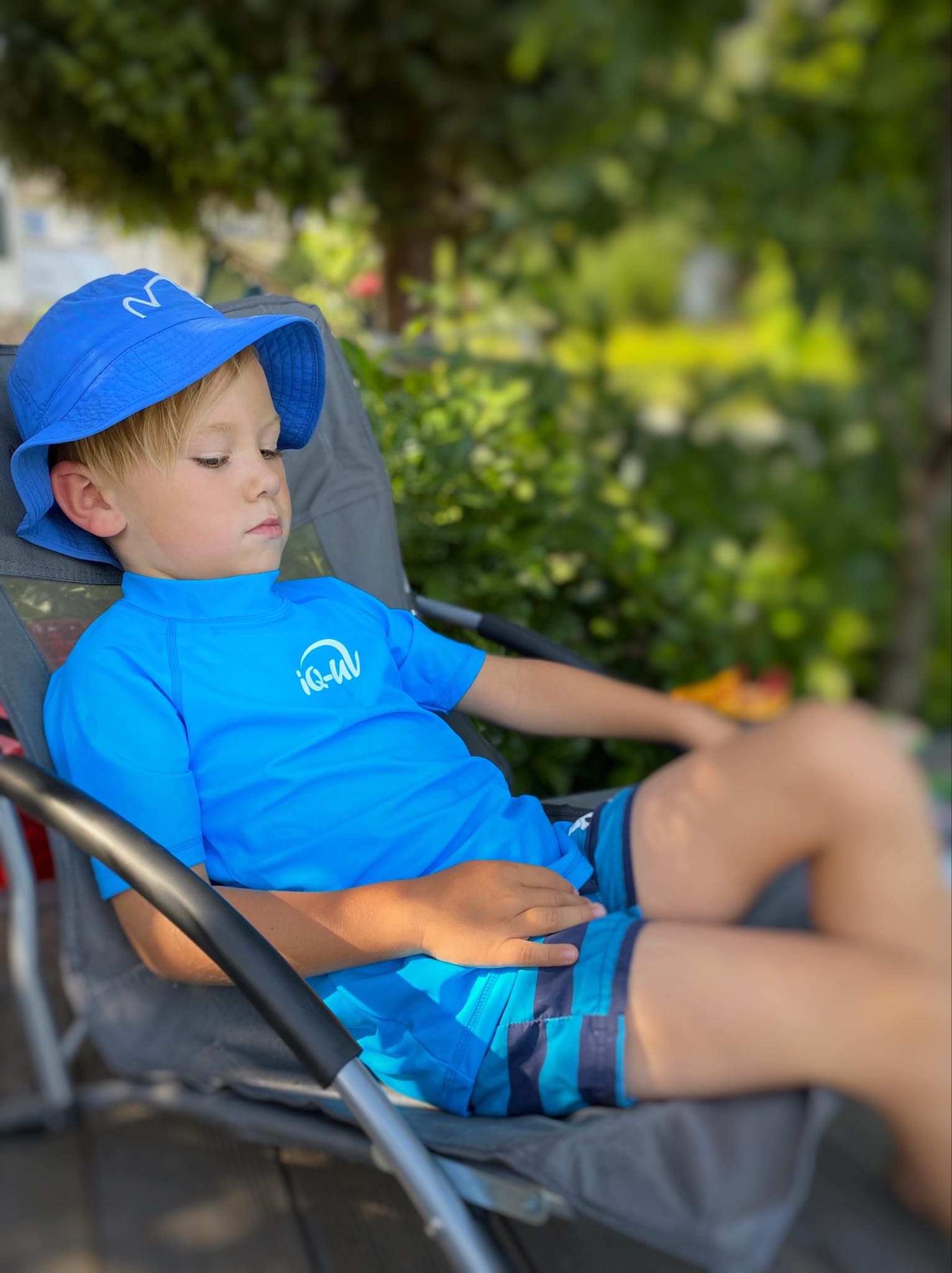 Kind sitzt in einem Gartenstuhl, trägt blaues Shirt und Hut mit iQ-UV-Logo. Kurze Hose. Sonnenbrille.