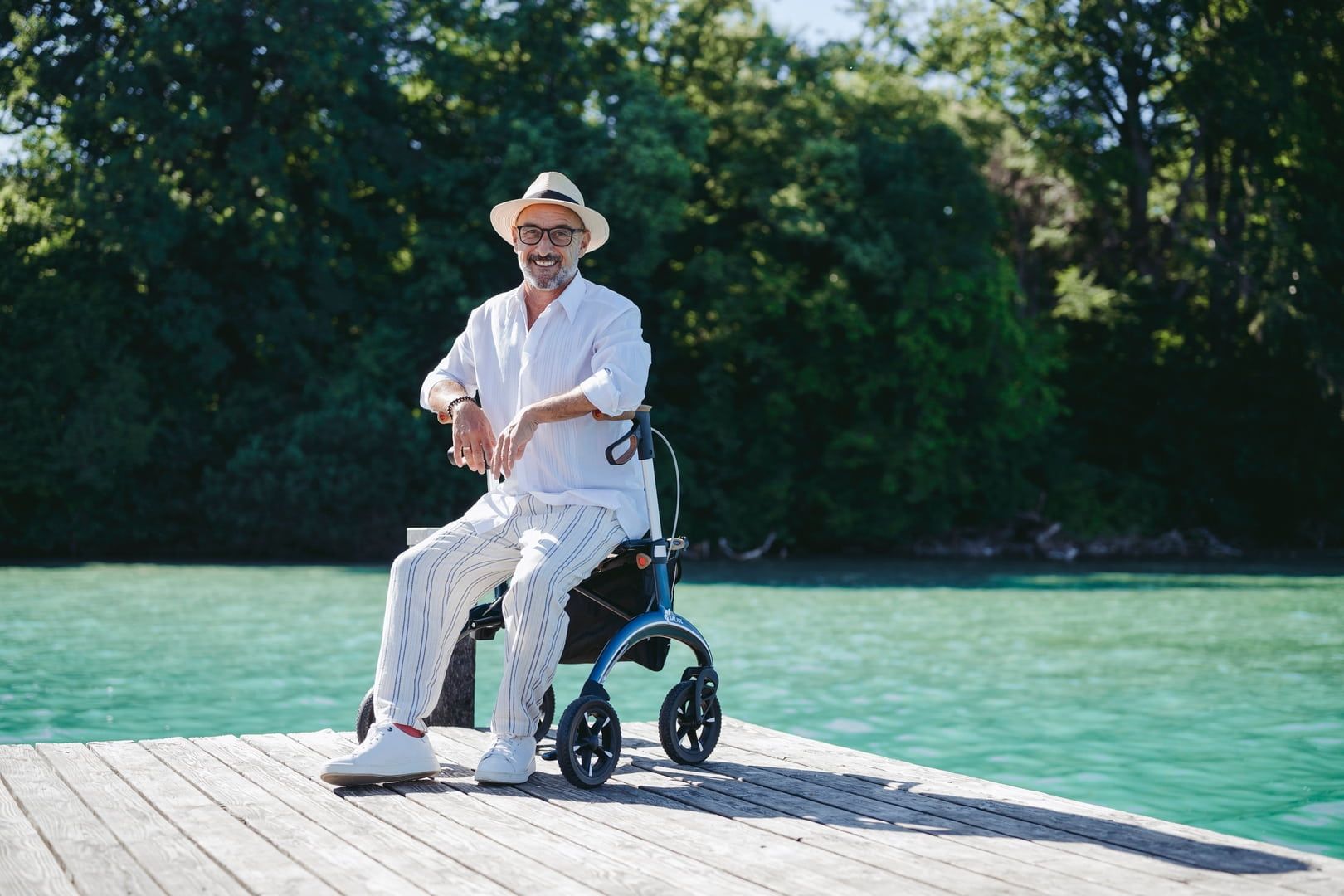 Ein Mann sitzt auf einem Steg neben einem blauen Rollator. Er trägt einen Hut und helle Kleidung.
