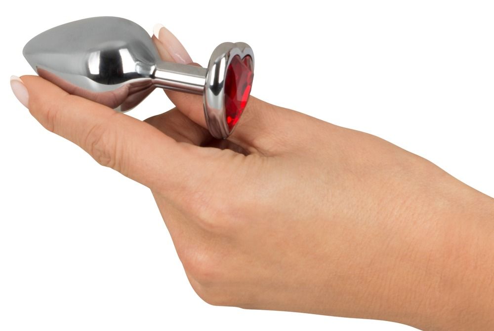 Hand hält Analplug mit herzförmigem, rotem Schmuckstein. Silberfarbenes Metall.