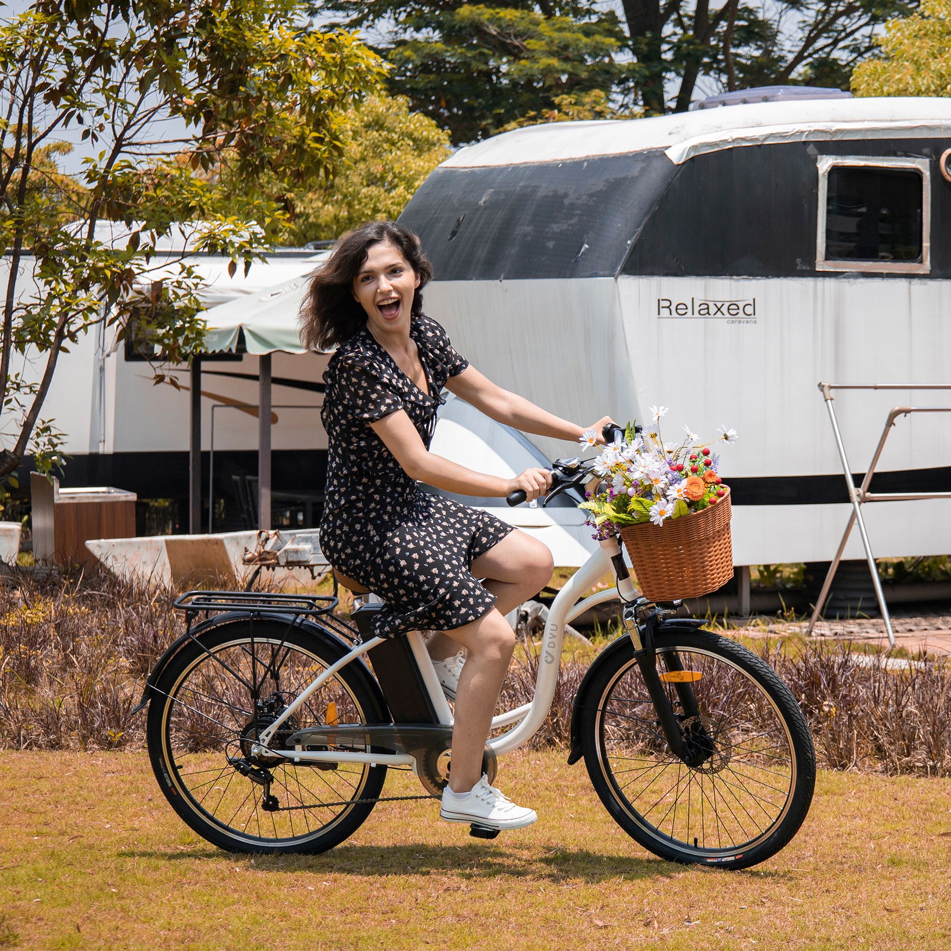 Frau auf weißem Fahrrad mit Korb, Blumen und schwarzem Rahmen. Im Hintergrund ein Gebäude.