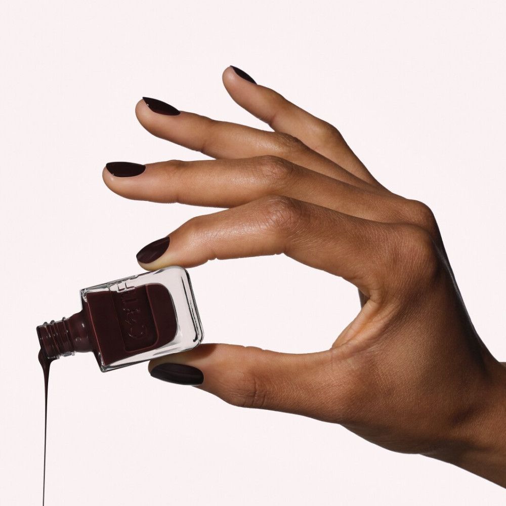 Dunkelbrauner Nagellack tropft aus einer Glasflasche, gehalten von einer Hand mit passenden Nägeln.