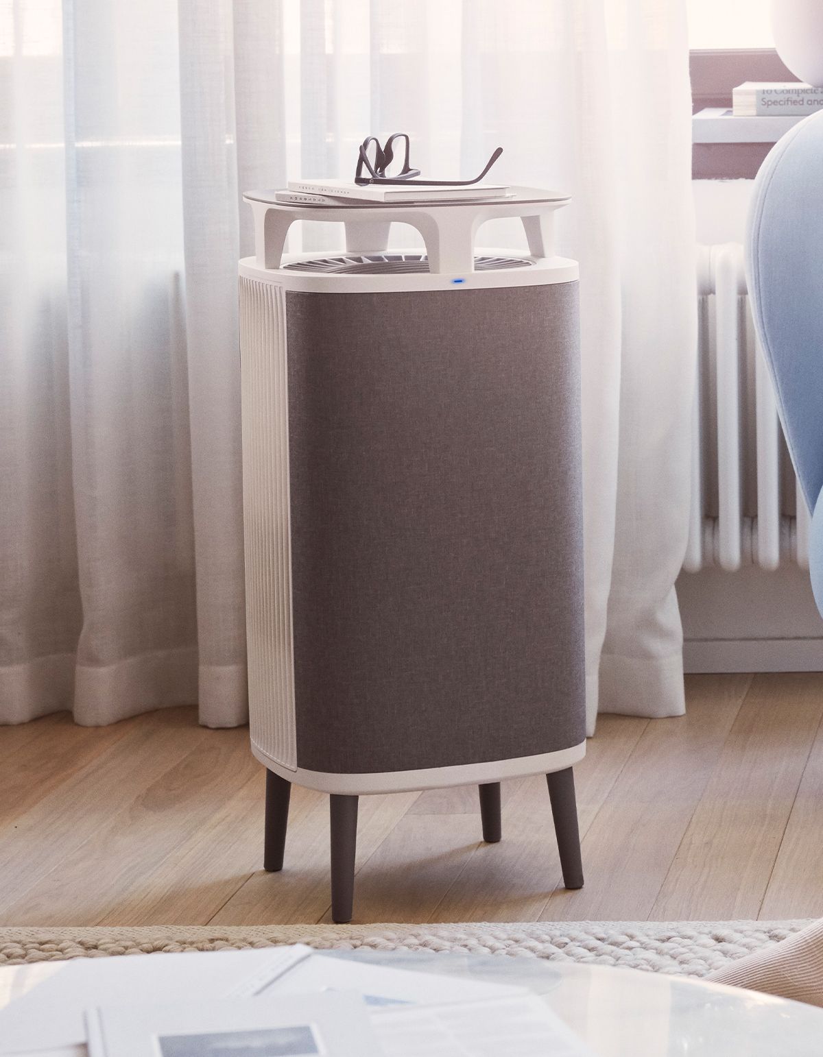 Luftreiniger in Wohnumgebung. Grauer Stoffbezug, weißer Rahmen, Beine. Brille und Notizen oben.