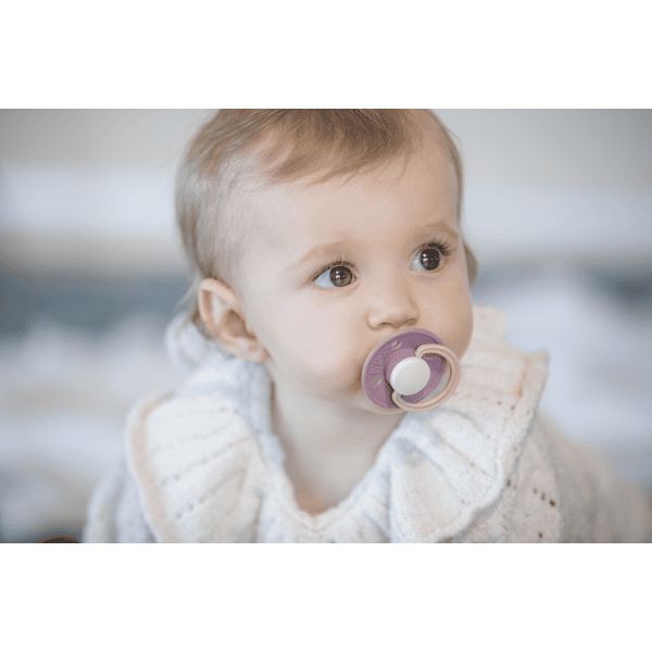 Ein Baby mit einem Schnuller im Mund. Der Schnuller hat einen runden Sauger und einen flachen Schild. Farbe: Lila.