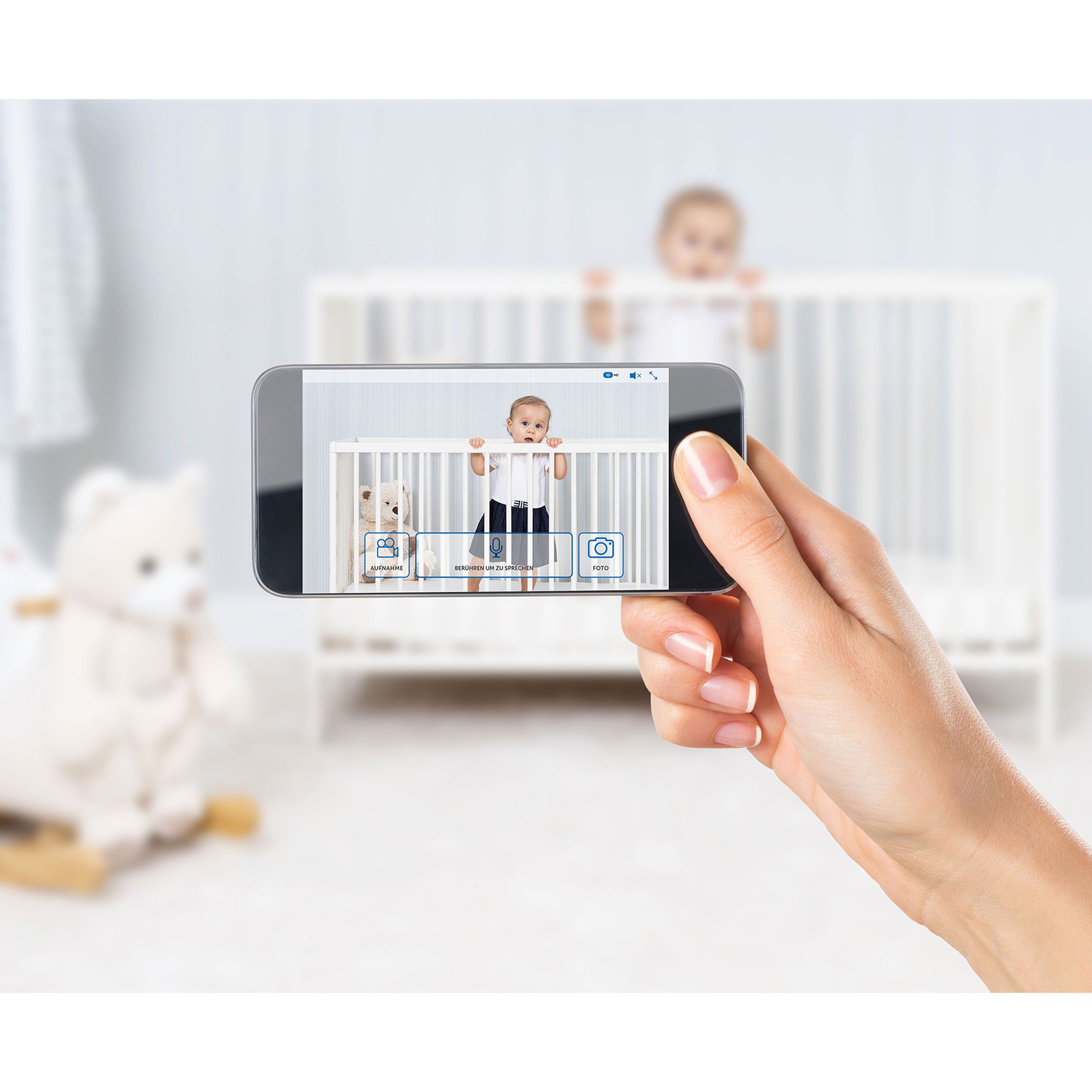Smartphone zeigt Baby in einem Kinderbett. Die Hand hält das Telefon. App-Symbole sind sichtbar.