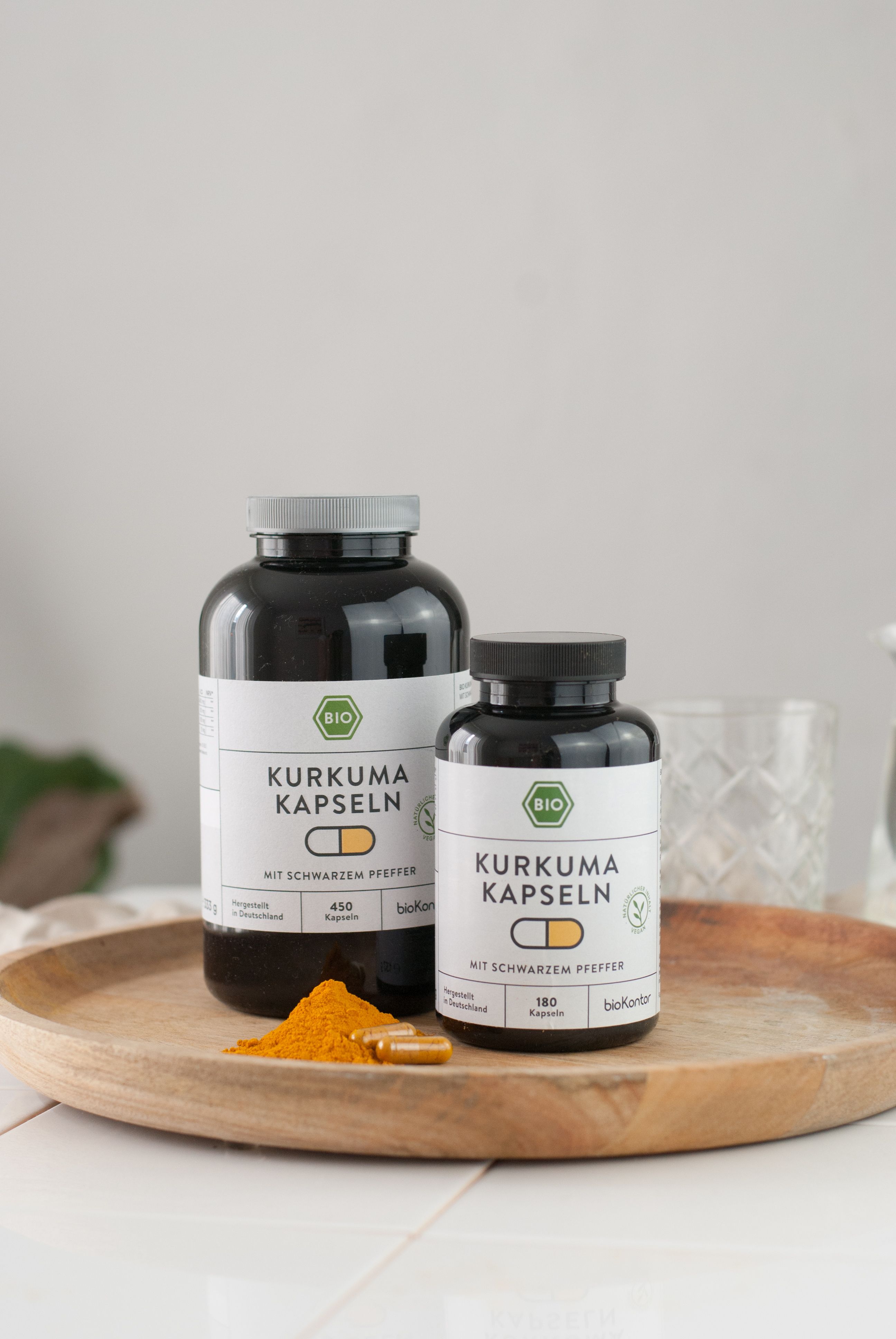 Zwei Flaschen Kurkuma Kapseln auf einem Holztablett. Eine kleine und eine große Flasche. Daneben Kurkumapulver und Kapseln.