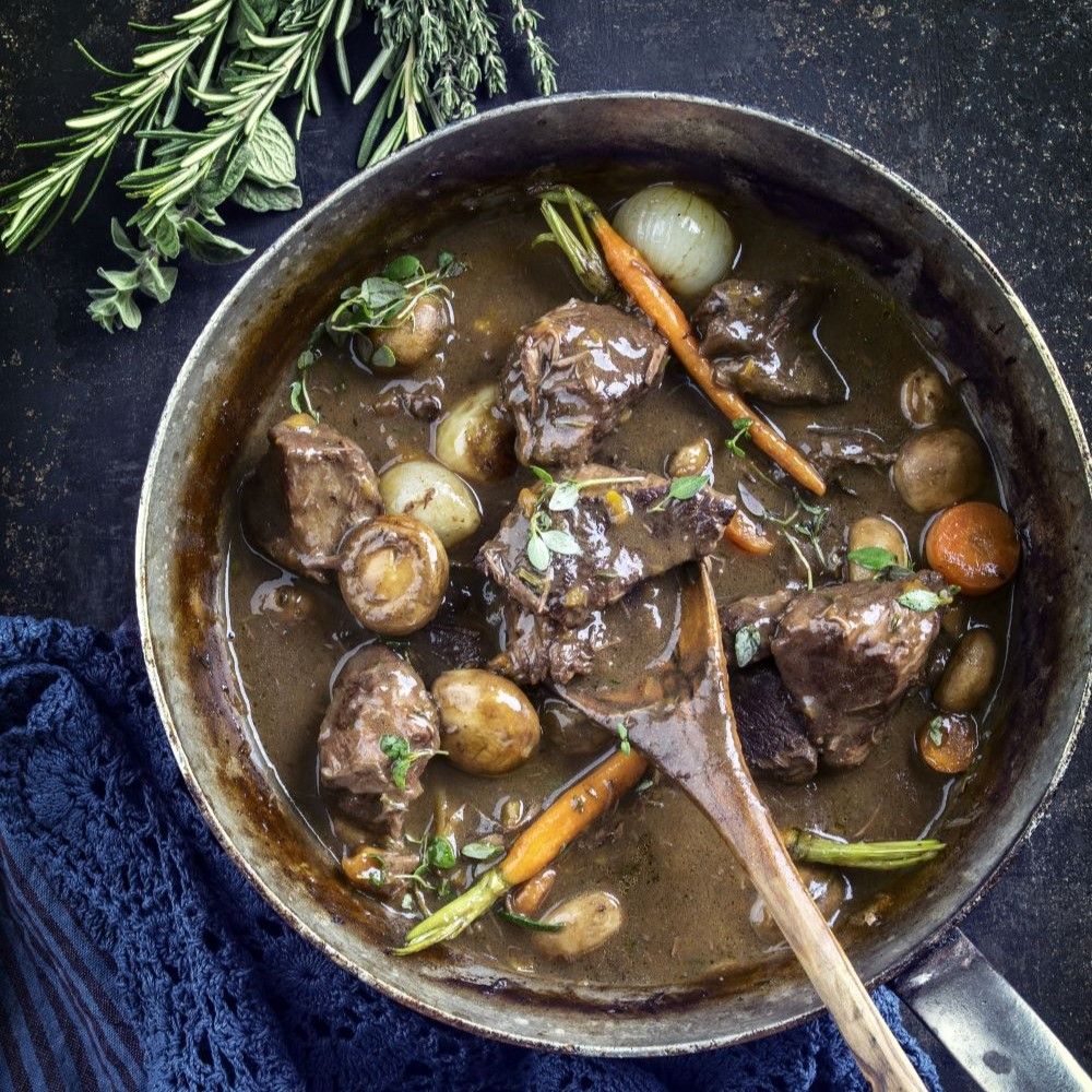 Eintopf in einer Pfanne. Enthält Fleisch, Gemüse und Kräuter. Holzkochlöffel im Eintopf. Blaue Serviette am Rand.