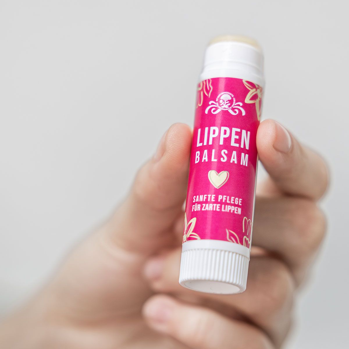 Hand hält Lippenbalsam-Stift. Pinkfarbene Hülle mit Aufschrift: LIPPEN BALSAM. Weißer Deckel. Sanfte Pflege für zarte Lippen.